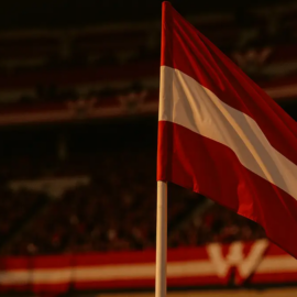 Austrian flag-coloured stadium stands with supporters, corner flag in sharp focus, evening floodlit atmosphere.