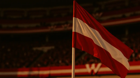 Austrian flag-coloured stadium stands with supporters, corner flag in sharp focus, evening floodlit atmosphere.