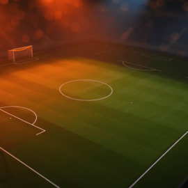 Floodlit football pitch at dusk with geometric white markings and orange-gold evening lighting creating dramatic shadows across emerald turf.