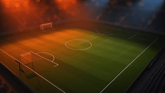Floodlit football pitch at dusk with geometric white markings and orange-gold evening lighting creating dramatic shadows across emerald turf.