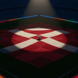 Aerial view of floodlit football pitch with red and white markings, ball on centre spot, empty stadium bowl beneath.