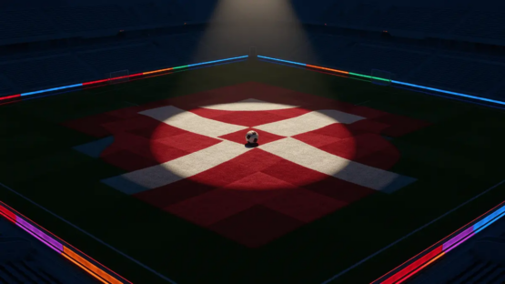 Aerial view of floodlit football pitch with red and white markings, ball on centre spot, empty stadium bowl beneath.