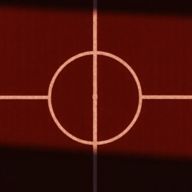 Overhead view of a floodlit football pitch with red grass and white markings at sunset.